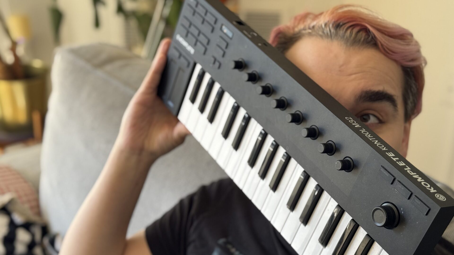 A person with faded pink hair holds a black midi keyboard in front of their face.